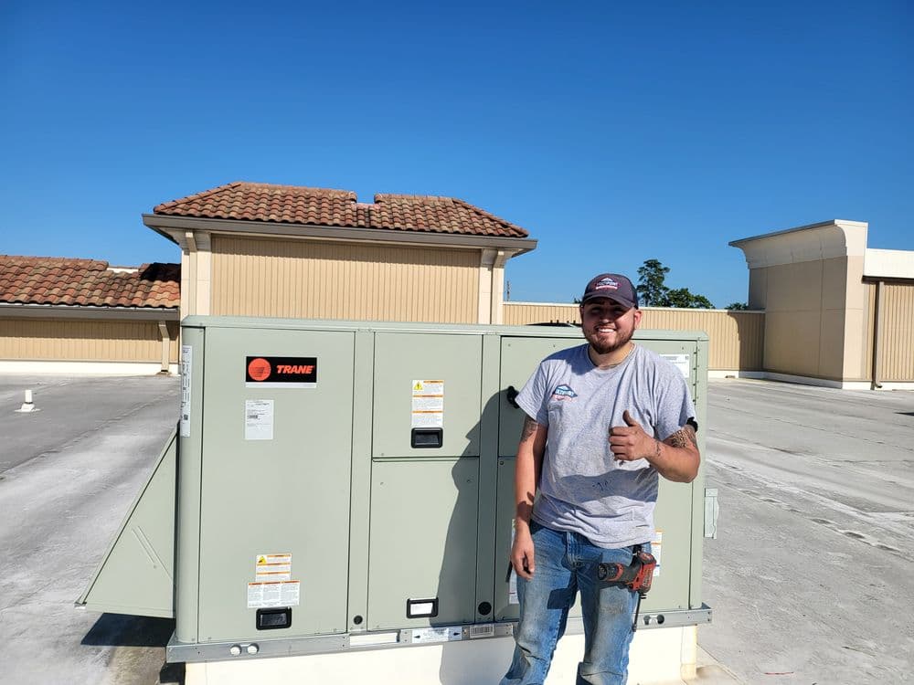 Technician standing next to rooftop HVAC unit by Trane under clear blue sky.