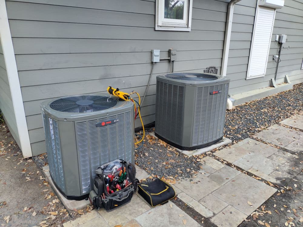 Two air conditioning units outside a gray house, with tools and equipment nearby.