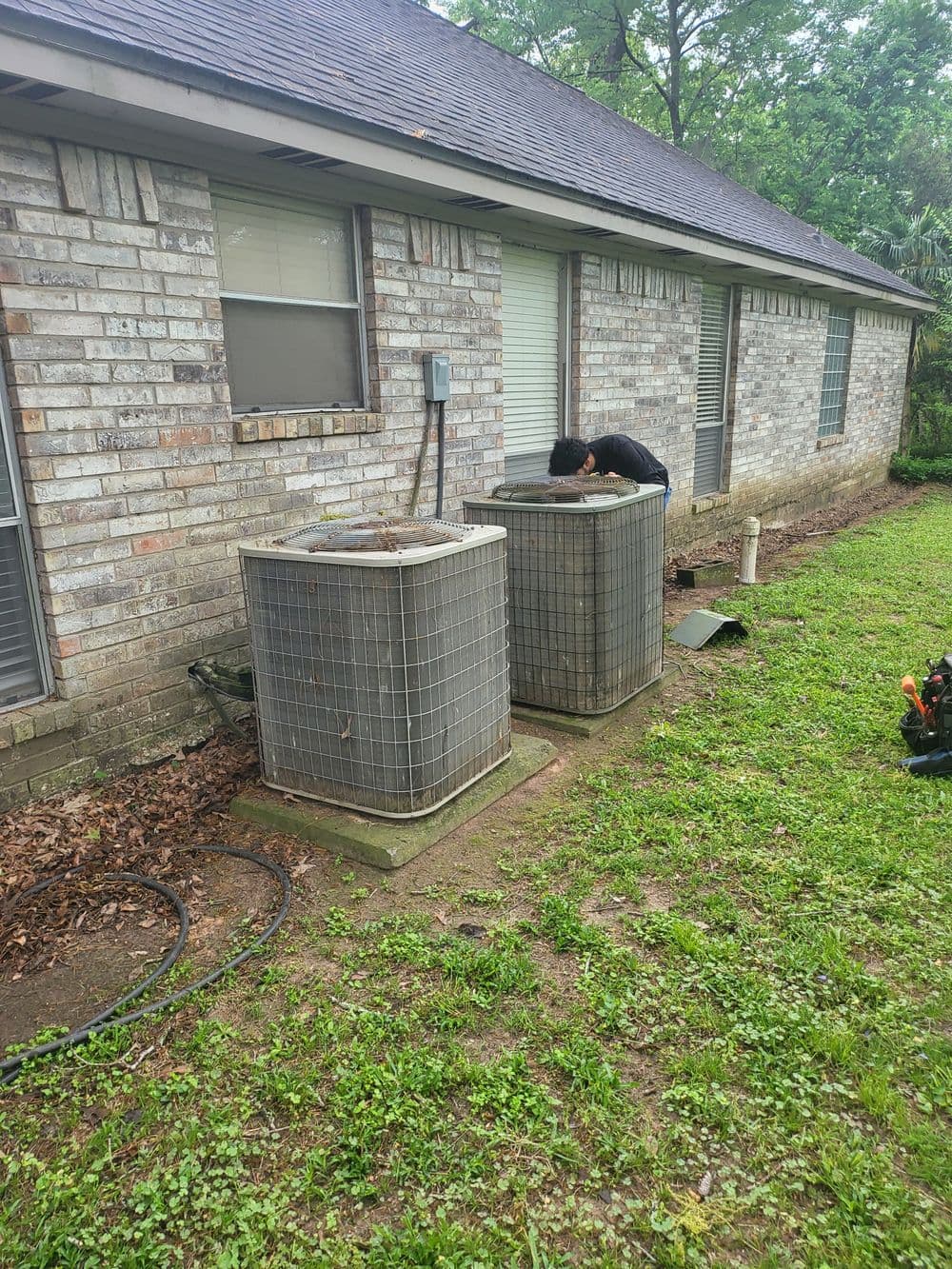 Technician servicing two outdoor HVAC units beside a brick house with greenery.
