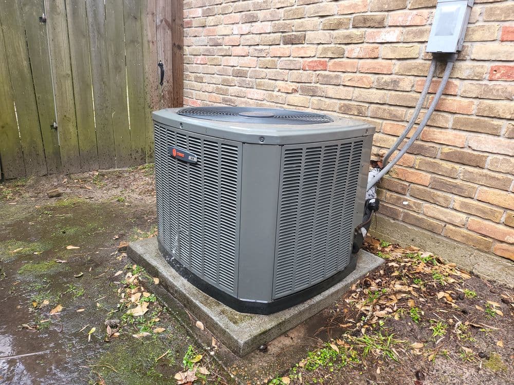 Gray outdoor air conditioning unit next to a brick wall and wooden fence.